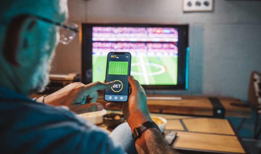Man looks at gambling app on a phone while watching live sport on TV