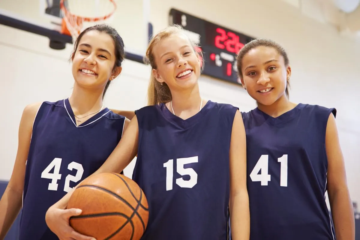 Three smiling young girls in purple basketball jerseys with one holding a basketball