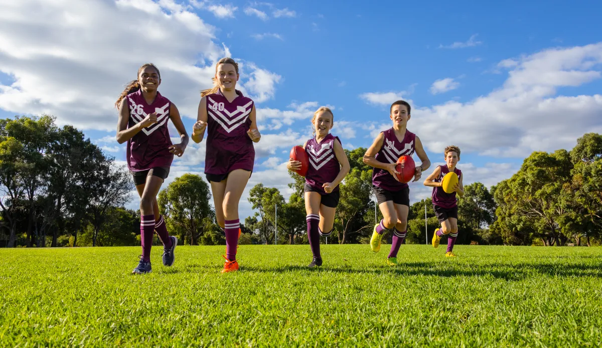 Australian childen and young people run across green grass playing Australian Rules football in purple and white uniforms