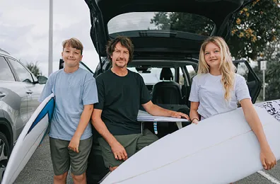 father and two children with surfboards behind a parked car with the boot open