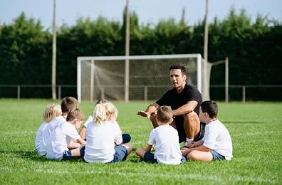 male coach talking to kids on a soccer field who are sitting in a circle