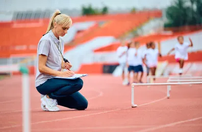 Athletics coach crouching down on athletics track with kids in the background jumping over hurdles