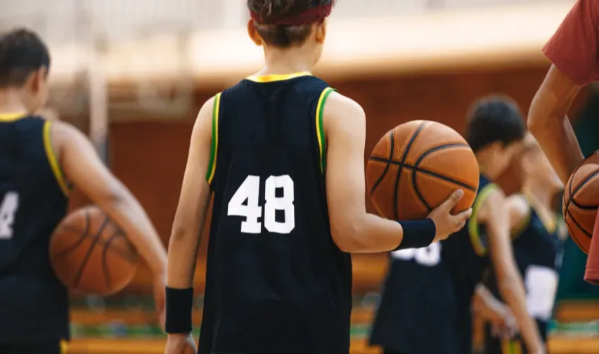 A young basketballer holds a basketball in hand with number 48 in white on the back of their black singlet