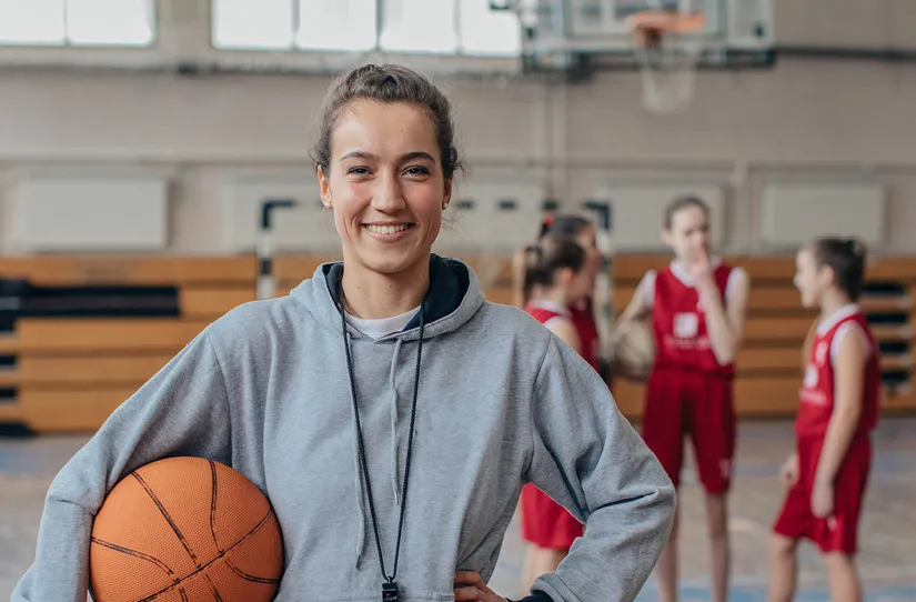 A female physical education teacher holds a basketball under one arm in a school gymnasium