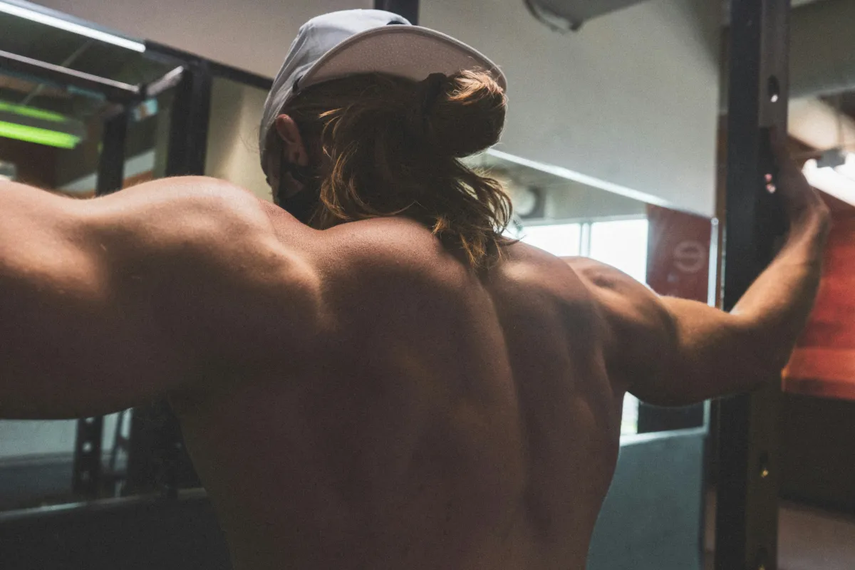 Man without shirt on and hat on backwards works out in a weights room